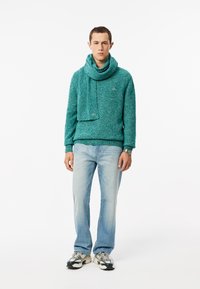 Maglione lavorato a maglia color turchese con sciarpa coordinata, entrambi con un design testurizzato. Indossato con jeans azzurri chiari e scarpe da ginnastica bianche.