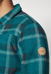 Tyrkysová a šedá kockovaná flísová košeľa na ramene s golierom a okrúhlym hnedým nášivom s nápisom "Icepeak Established 1996" na hornej časti rukáva.