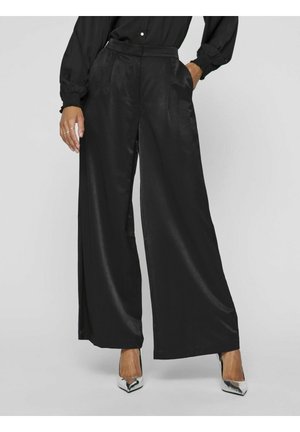 Pantalones amplios negros hechos de una tela suave y brillante. Presentan una cintura alta, bolsillos laterales y un diseño fluido. Combinados con tacones plateados.