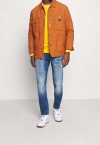 Orange kordjacka med en gul tröja under, tillsammans med slitna blå jeans och vita sneakers, med en diskret logopatch.