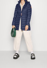 Laivasto sininen puhvitakki, jossa on huppu ja pystysaumat, yhdistettynä kermaisiin leveälahkeisiin housuihin, mustiin kohotettuihin sandaaleihin ja vihreään käsilaukkuun.