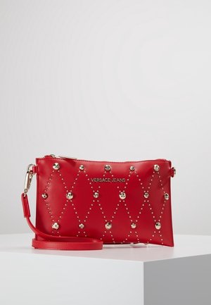 Petite pochette de poignet Versace Jeans rouge avec des motifs de clous en forme de losange, une quincaillerie dorée et une sangle de poignet amovible sur une surface blanche.
