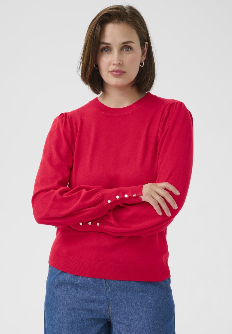 Red pullover sweater with long, puffed sleeves and button accents at the cuffs. Smooth texture and classic crew neckline. Denim jeans visible.