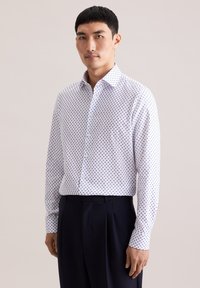 Camicia con bottoni bianca a pois blu, maniche lunghe e colletto regolare. Abbinata a pantaloni blu scuro.