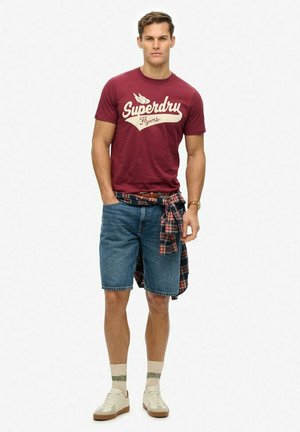 Bordeaux bomulds T-shirt med "Superdry Flyers" print, blå denimshorts, ternet skjorte bundet omkring taljen, beige sneakers og creme-grønne sokker.