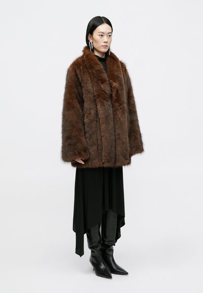 Abrigo de piel marrón con un collar amplio, que presenta una textura suave. Llevado sobre un vestido negro asimétrico y botas altas de tacón negro.