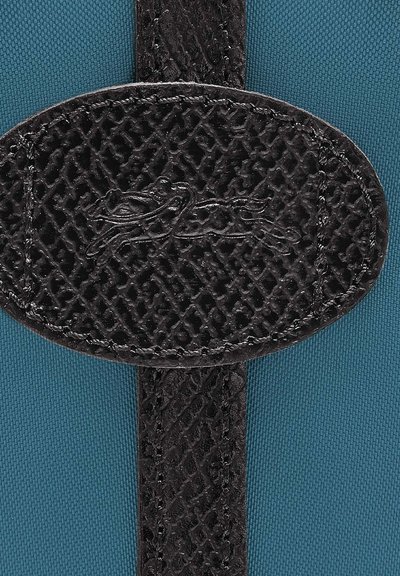 Texturiertes schwarzes Lederpatch mit geprägtem Design auf einem blauen Stoffhintergrund, ausgestattet mit einem stabilen Riemen und runder Form.