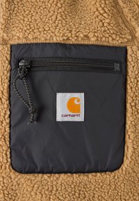 Bruine fleece stof met een zwarte nylon zak. Voorzien van een ritssluiting en een wit Carhartt logo patch. Gestructureerd oppervlak, duurzaam ontwerp.