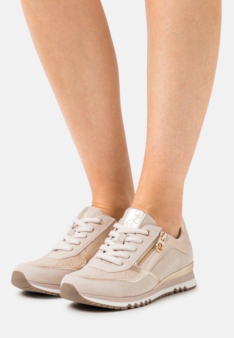 Sneakers in suede beige con pannelli laterali testurizzati, accenti dorati, logo riflettente e suola bianca. Presentano una chiusura con lacci frontale e una cerniera laterale.
