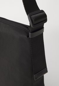 Černá textilní crossbody taška s hladkou texturou, nastavitelným popruhem a obdélníkovým tvarem. Obsahuje plastovou sponu na hardware.