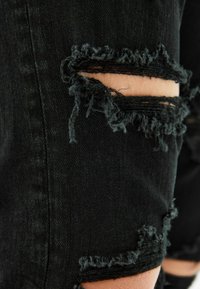 Svarta denimjeans med fransade sprickor vid knäna. Strukturerad tyg med en urblekt finish och lösa trådar vid sprickkanterna.