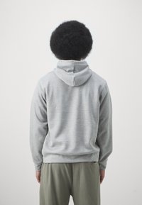 Felpa grigia in cotone con cappuccio regolabile, caratterizzata da una consistenza morbida e un fit rilassato. Polsini e orlo a coste ben visibili. I pantaloni sono di colore verde oliva.