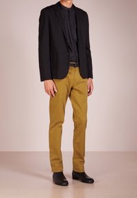Czarna marynarka na czarnej koszuli, zestawiona z musztardowymi spodniami i czarnymi skórzanymi butami. Gładki materiał i dopasowany krój.