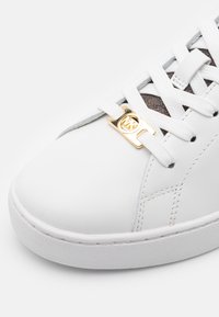 Baskets blanches en cuir avec lacets blancs, breloque dorée avec le logo "MK" sur les lacets, et tissu texturé marron près de la languette.