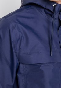Navy hooded jacket made from smooth, water-resistant material. Features drawstrings, a front pocket, and a subtle grid pattern.