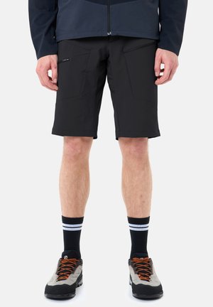 Uomo che indossa pantaloncini neri fino a metà coscia con tasche laterali con zip, calzini neri con due strisce bianche e scarpe da trekking grigie con lacci arancioni.