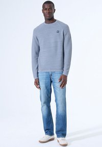 Sudadera de textura gris claro con cuello redondo y pequeño logo, combinada con jeans rectos azules y zapatillas blancas.