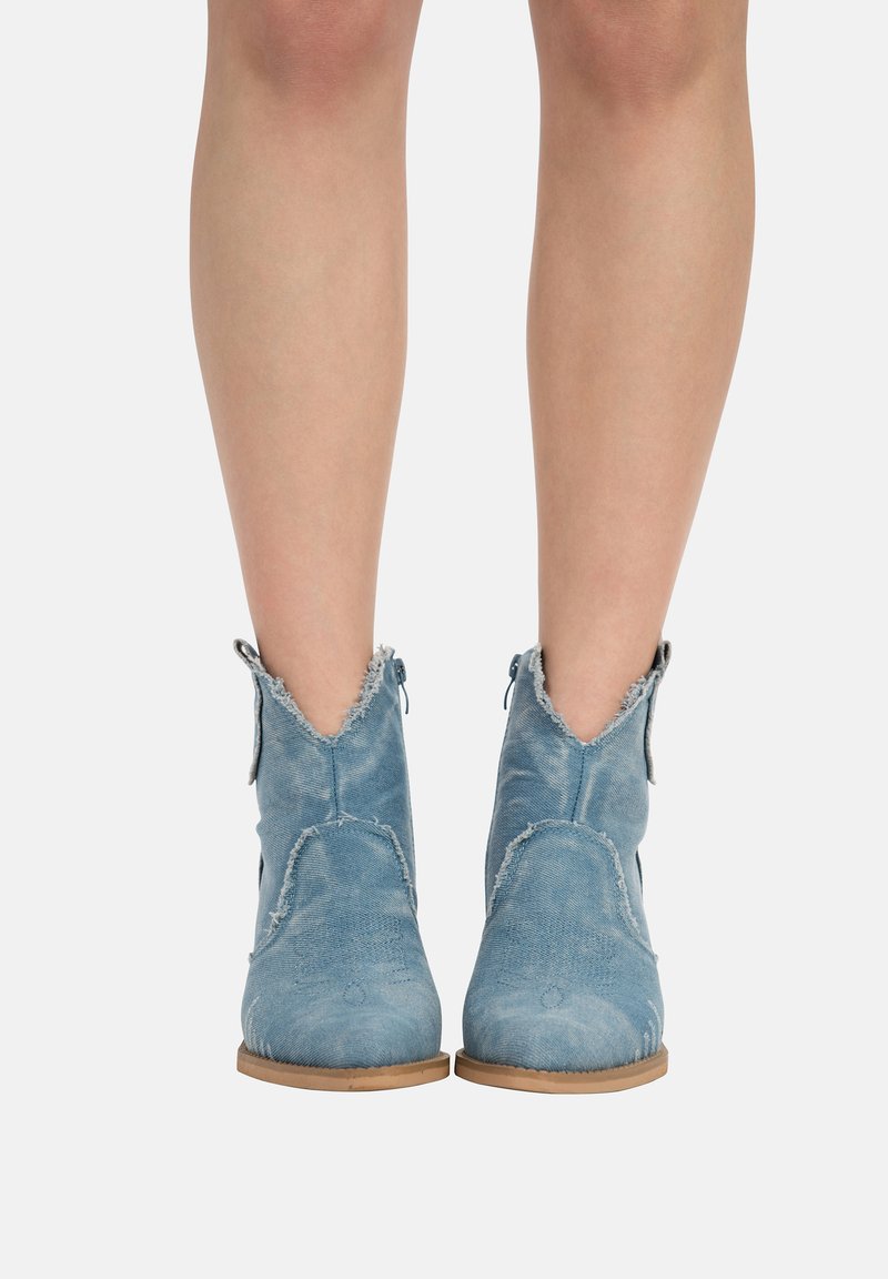 Botas de tobillo azules con una textura de mezclilla desgastada, que presentan un diseño en forma de V, bordes deshilachados y una suela de color marrón claro.