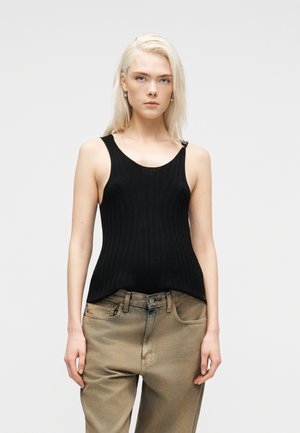 Ung kvinne med blondt hår iført svart ermeløs ribbestrikket topp og løse beige jeans, stående mot en ensfarget lys bakgrunn.