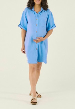 Femme enceinte portant une robe bleue boutonnée et des sandales, souriante, une main posée sur son ventre, sur un fond uni.