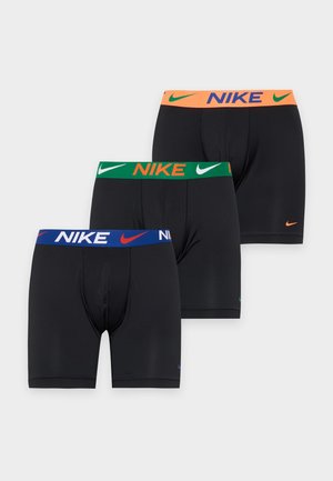 Un mannequin masculin se tient devant un fond blanc uni, vêtu d’un boxer Nike noir à ceinture bleue.