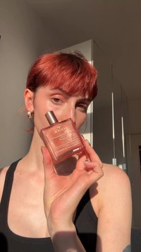 Persona con cabello corto y rojo sostiene un frasco cuadrado de perfume rosa con brillo etiquetado "NUXE" cerca de su rostro a la luz natural.