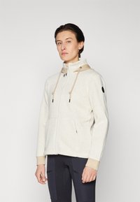 Cream herringbone zip-up jacket with a beige hood, adjustable drawstrings, front pockets, and black logo detail on sleeve.