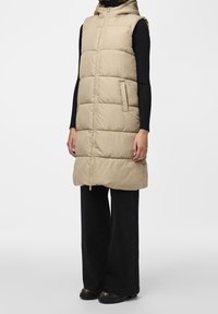 Beige vycpaná bezrukávová vesta s kapucí, předním zipem a bočními kapsami, nošená přes černý žebrovaný svetr a široké černé kalhoty.