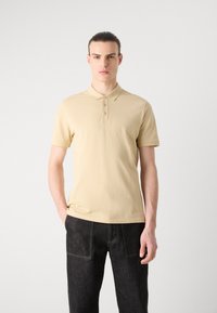 Beige pólóing puha textúrával, három gombos gallérral és diszkrét hímezett logóval a mellrészen, sötét farmernadrággal párosítva.