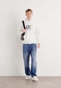 Hoodie blanc avec un logo "CK" multicolore, jeans bleus et baskets blanches. Le mannequin porte un sac à bandoulière noir. Fond uni.