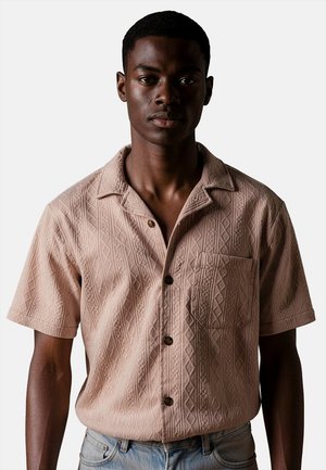 Giovane uomo con una camicia rosa chiaro a maniche corte con motivi a diamante testurizzati e una tasca sul petto, abbinata a jeans azzurri chiari.