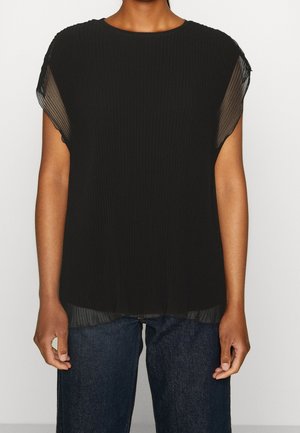 Blouse noire à manches courtes plissée avec manches transparentes superposées portée avec un jean bleu foncé sur une personne debout.