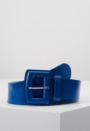 Blauwe leren riem met grote vierkante gesp en zilveren tonguegesp, opgerold en geplaatst op een witte ondergrond tegen een lichtgrijze achtergrond.