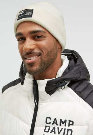 Padded jacket in white and black with a high collar, featuring a zip closure and "CAMP DAVID" logo. Accompanied by a cream beanie.