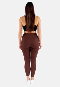 Brązowe legginsy wykonane z elastycznego materiału, z wysokim stanem i dopasowanym krojem, zestawione z czarną bluzką bez rękawów i białymi trampkami.