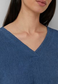 Femme portant un haut V bleu texturé, mettant en valeur le cou, la clavicule et le bas du visage avec les lèvres closes.