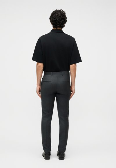 Άνδρας με σγουρά μαλλιά, φοράει μαύρο κοντομάνικο πουκάμισο και εφαρμοστά σκούρα γκρι παντελόνια, στέκεται με την πλάτη στο απλό φόντο.