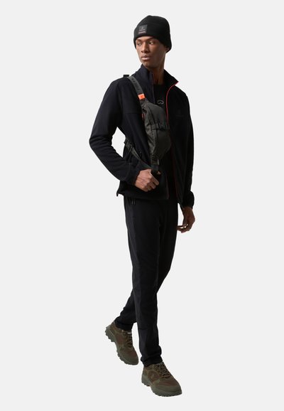 Sort fleece zip-jakke med røde detaljer, slim-fit sorte bukser og olivengrønne vandresko. Sort crossbody taske og strikket beanie.