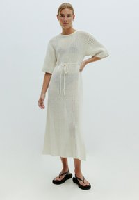 Weißes gestricktes Kleid mit kurzen Ärmeln, Kordelzug in der Taille und lockerer Passform, mit offenem Gewebemuster; das Model trägt schwarze Flip-Flops.