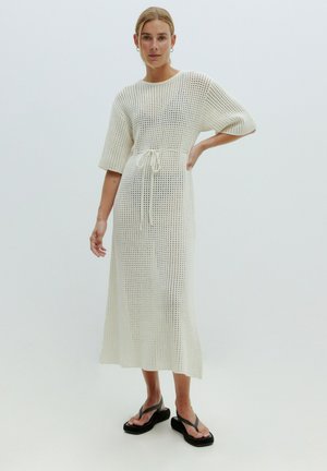Weißes gestricktes Kleid mit kurzen Ärmeln, Kordelzug in der Taille und lockerer Passform, mit offenem Gewebemuster; das Model trägt schwarze Flip-Flops.
