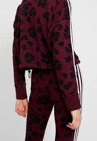 Sudadera recortada color burdeos con estampado de leopardo negro, que presenta rayas blancas en los lados de las mangas. Combinada con leggings ajustados a juego.