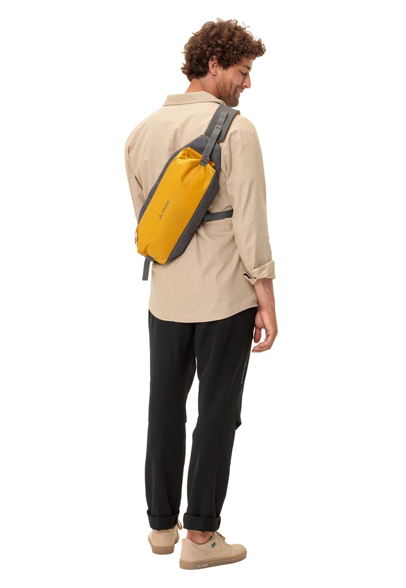 Zaino a tracolla giallo e grigio con una finitura testurizzata, una sola cinghia e una chiusura con zip, indossato sulla schiena di una persona in abbigliamento casual.