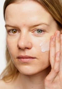 Crème hydratante appliquée sur une peau claire, une crème rose pâle s'étend le long de la joue ; les caractéristiques marquantes incluent une peau texturée et une diversité de couleurs d'yeux.