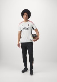 Vit tröja med rosa och svarta detaljer, med "QATAR AIRWAYS"-logotyp. Svarta byxor med orange detaljer. Svart fotboll i handen.