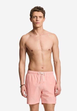Young man with short curly hair wearing pink and white striped swim shorts with drawstring, standing against a plain white background.
