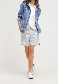 Jeansowa kurtka z czerwoną naszywką w kształcie gwiazdy, biała koszula z guzikami, wzorzyste niebieskie szorty i białe sneakersy. Casualowy strój z warstwami tekstur.