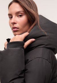 Junge Frau passt den hohen Kragen einer schwarzen Daunenjacke an und schaut über ihre Schulter vor einem neutralen Hintergrund.