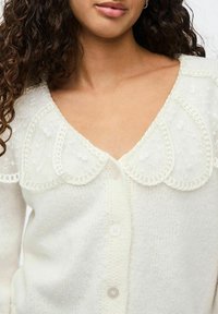 Vit stickad cardigan med en scalloped spetskrage och delikata detaljer, med knäppning och mjuk textur.