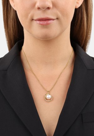 Collar de oro con un colgante de perla round, que presenta un diseño circular y pequeños acentos transparentes, llevado sobre un blazer negro.