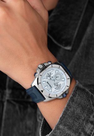Montre argentée avec un cadran blanc texturé, des accents bleus et un bracelet en silicone bleu. Comprend des marqueurs d'heure audacieux et des sous-cadrans chronographes.